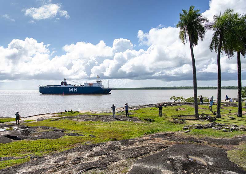 A ship, carrying NASA’s James Webb Space Telescope as cargo, arrives. In the foreground there are several people on land standing by taking photos.