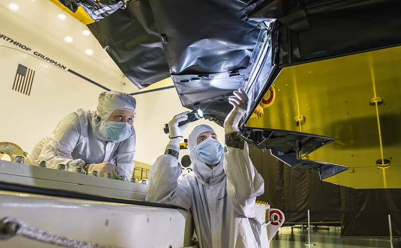 Two people wearing cleanroom suits, masks, and gloves inspect part of the Webb's primary mirror using a flashlight.