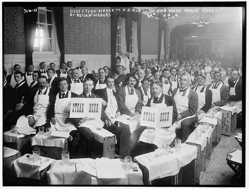 A black and white photo of a crowd of men in a restaurant in aprons holding signs that say STEAK and BEER.