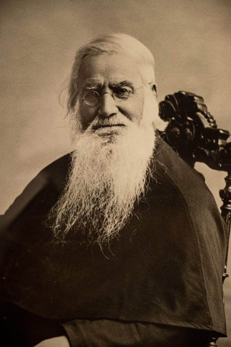 Sepia portrait of a man with gray hair and a long gray beard sits in a chair. He is wearing black clerical robes.