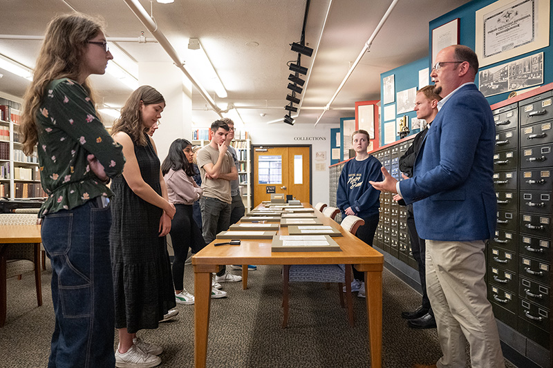 A group of men and women stand around a table in the middle of a room with brown mats with documents on them. They are surrounded by card catalogs and library shelves.