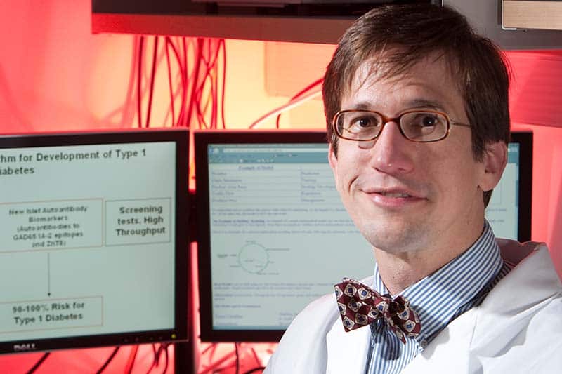 Schnell, in bow tie and white lab coat, in front of two computers with models and equations on the screens. The room has a red glow.