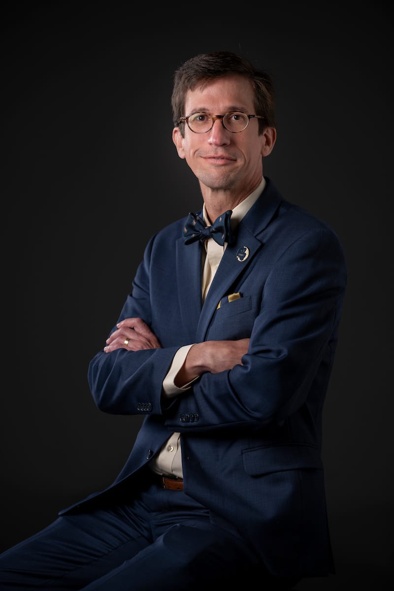 Schnell, wearing a bow tie and a navy suite, sits with his arms crossed in front of a dark background.