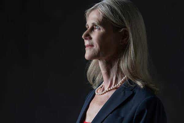 Side portrait of a woman against a dark backdrop. She is wearing a blazer over a red shirt and a pearl necklace. 