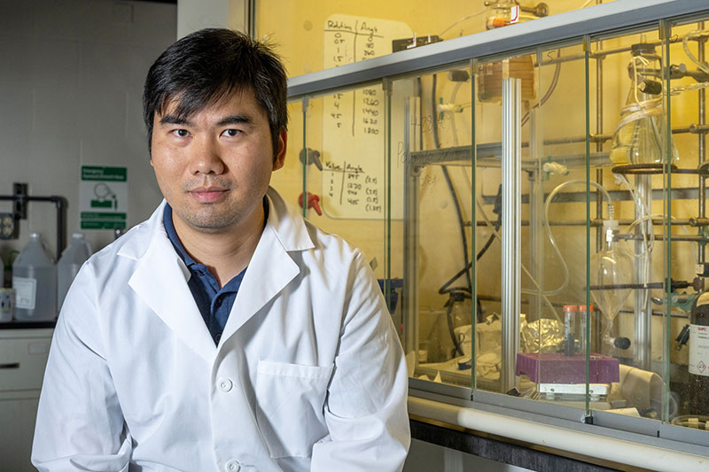 A man poses for a portrait in front of a glassed-in space filled with laboratory glassware and equipment.