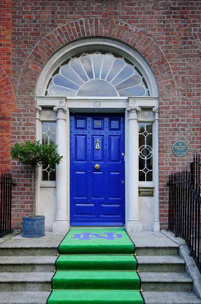 A brick building with a blue door and green Notre Dame rug on the front steps