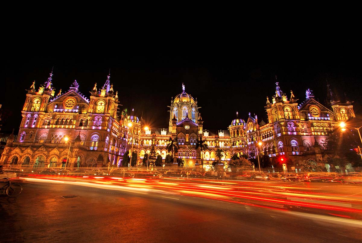 The ornate building is lit up with gold and purple light at night, with red blurs of lights from cars driving by in front of the building.