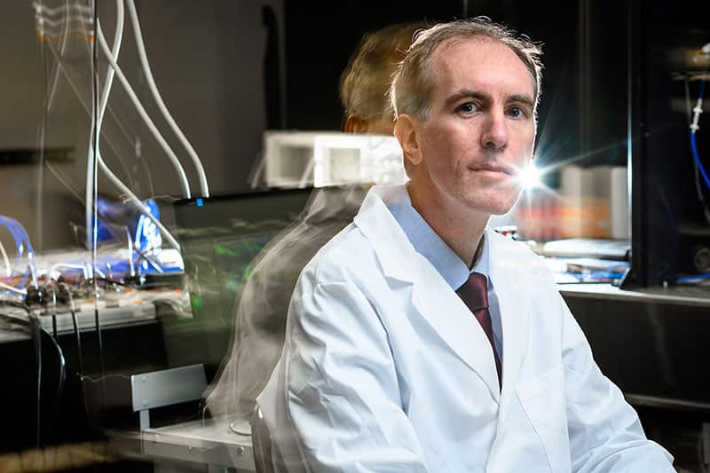 A man poses for a photo in a lab. Lights and reflections shine behind him.