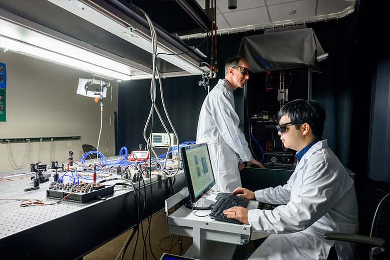 Two people wearing lab coats and eye protection glasses. One sits at a computer and another stands near lab equipment.