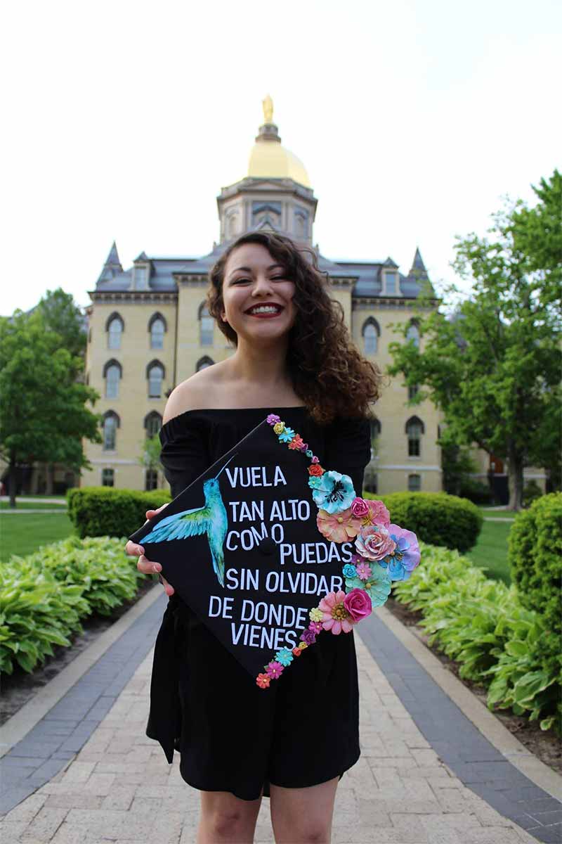 A female student wears a black dress, standing in front of the Golden Dome, holding a decorated graduation cap that says “Fly as high as you can without forgetting where you come from” in Spanish. There is a hummingbird and colorful flowers that border the cap.
