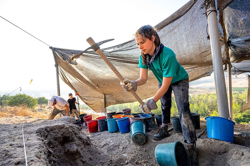 A woman surrounded by buckets uses a pickaxe to break up dirt.