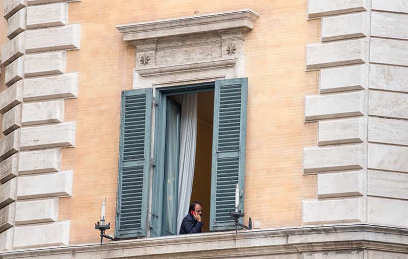 A man talks on the phone while sitting next to a balcony window.