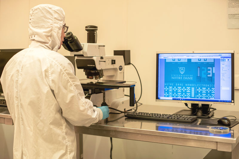 A person in a hazmat suit looks into a microscope. Next to that person is a computer screen that shows a close up of a microchip.