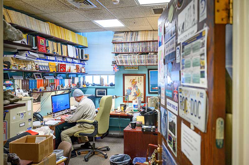 The inside of an office a man sits at a desk surrounded by hundreds of books and documents.