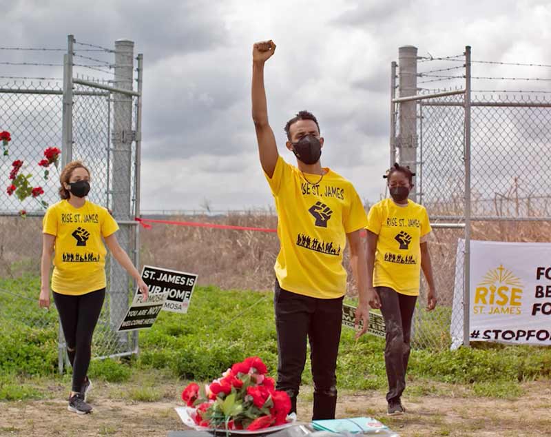 Three people stand in front of a tall chain linked fence. One person holds their fist in the air.