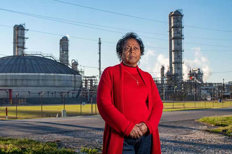 A woman wearing a red sweater and cross necklace stands in front of a chemical plant looking off camera.