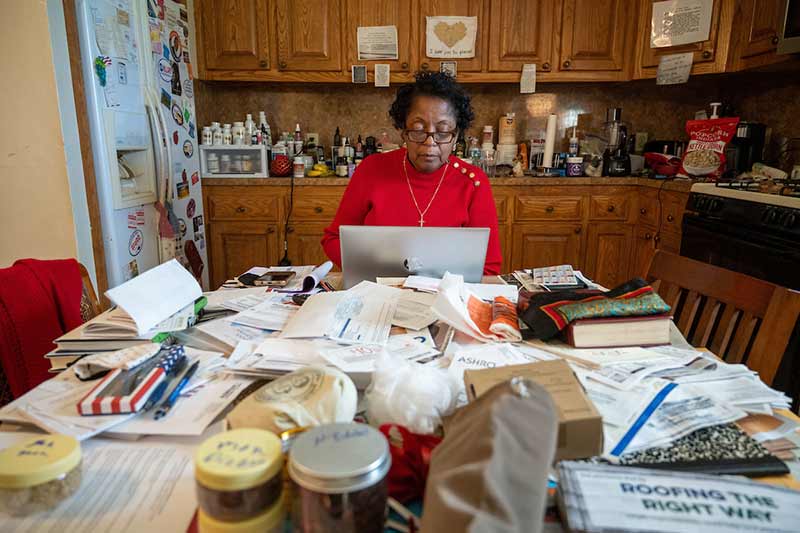 A woman works on her laptop at a cluttered kitchen table.