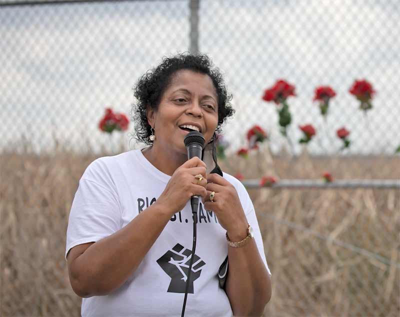 Outside a woman speaks into a microphone. Blurred out behind her is a tall chain linked fence with red flowers.