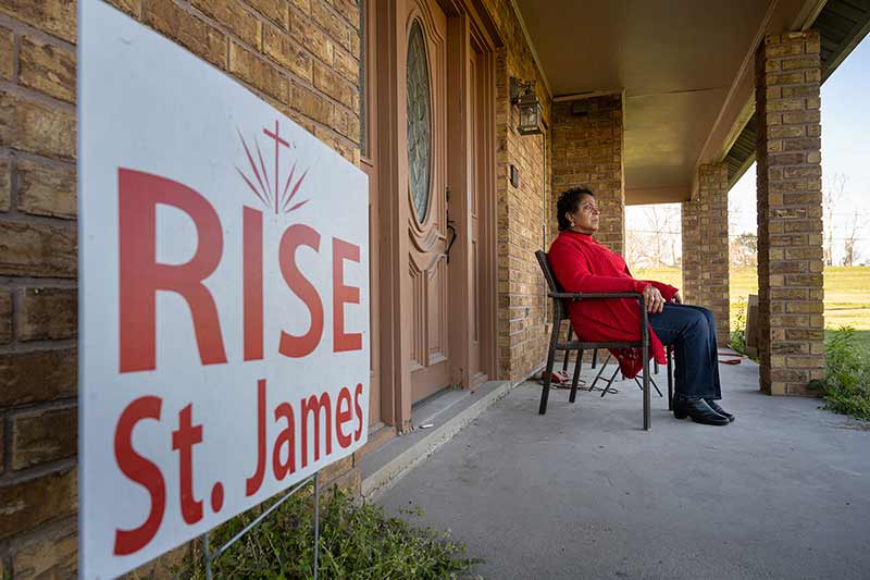 A woman sits in a chair on her front porch. A 'Rise St. James' yard sign in the foreground.