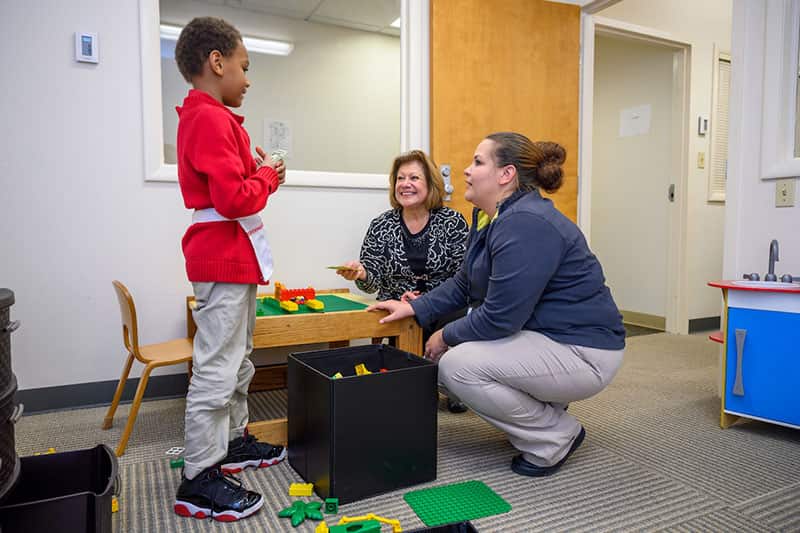 A young boy plays with colorful Legos while talking to two women.