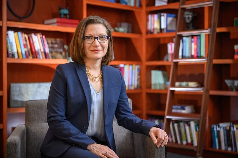 A woman wearing a blue blazer and frames sits in a sofa chair. Book shelves stand behind her in the background.