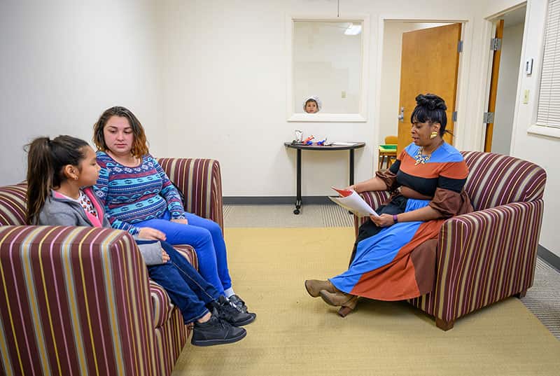 A mother and her daughter both sit on a couch while talking to another woman sitting on a sofa chair across from them. A little boy pokes his head up from another room in the background.