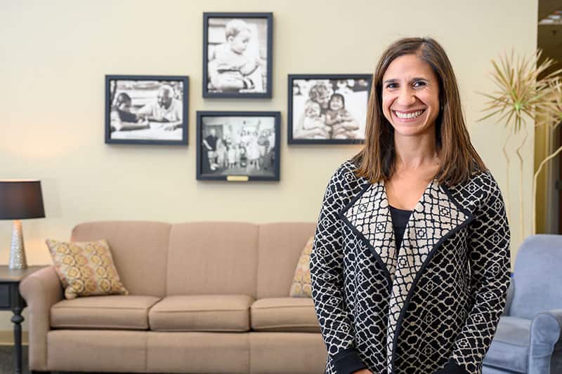 A woman wearing a long cardigan with black and white patterns stands in a room that resembles a living room. There are framed black and white photos of children on the wall in the background.