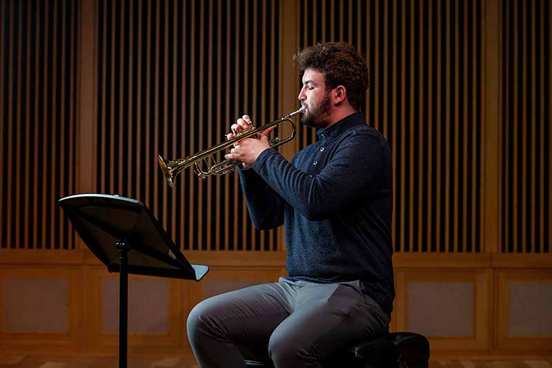 A male student plays a trumpet sitting on a stage with a stand for sheet music in front of him.