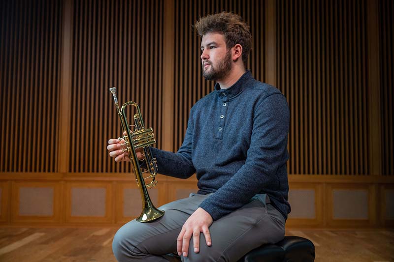 A male student sits with his trumpet resting on his knee on a stage.