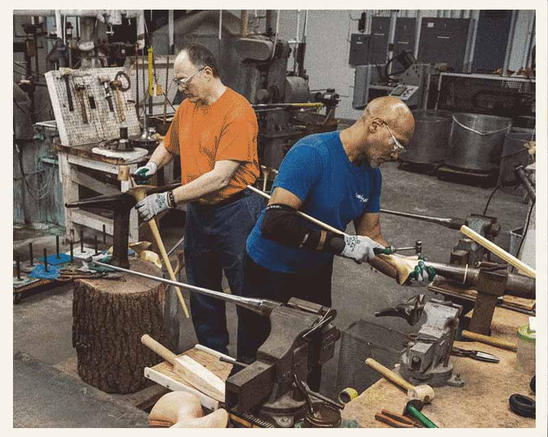 A current color photo of two men, one White and one Black, shape bells by hand using hammers and mandrels.