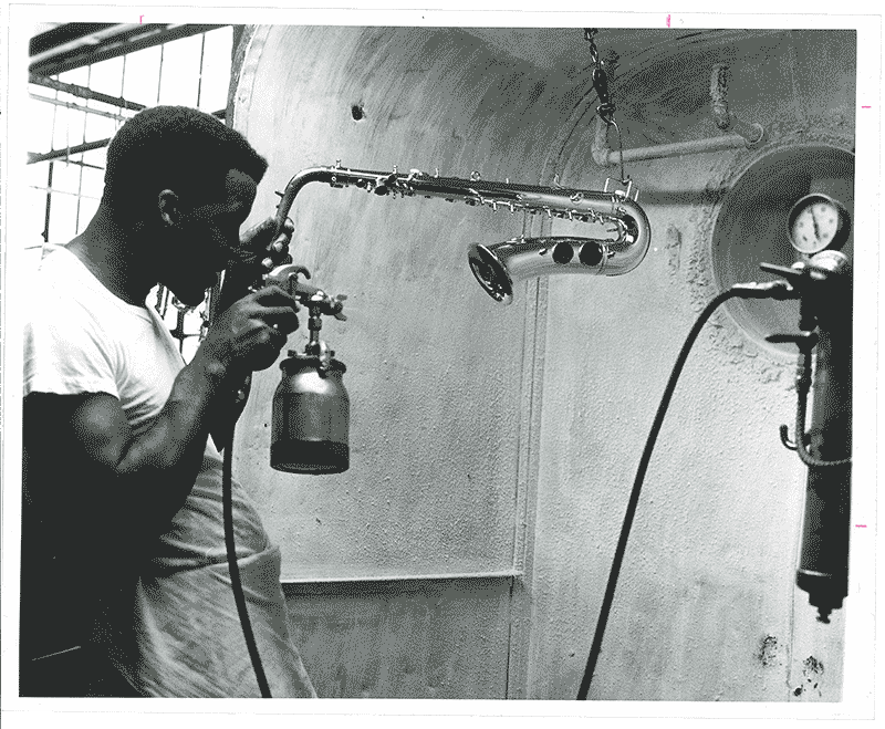A black and white archival photo of a Black man using a paint sprayer on a saxophone.