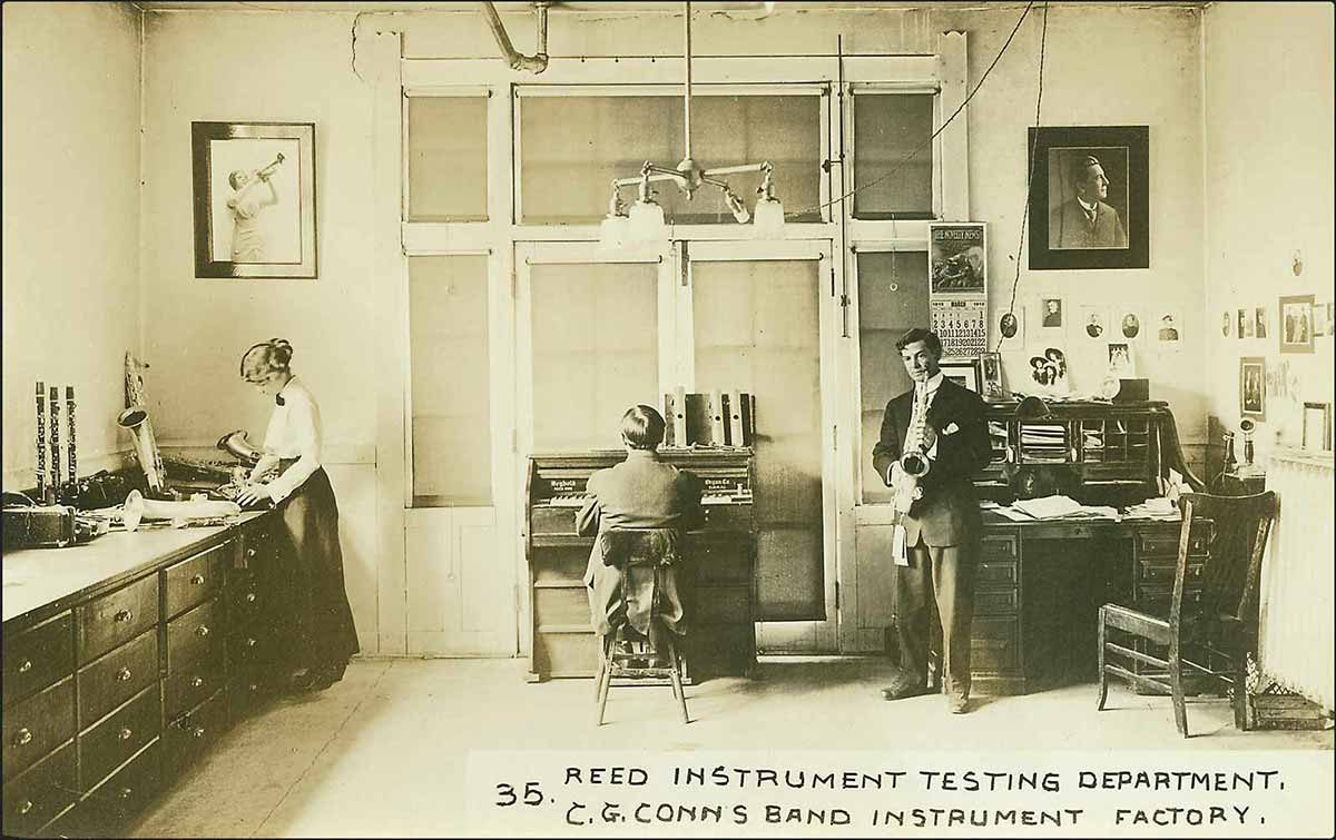 A yellowish archival photo of two men and a woman in a room. One man sits at a piano with his back to the camera, and the other stands and holds a saxophone. The woman is standing and working at a counter on a saxophone.