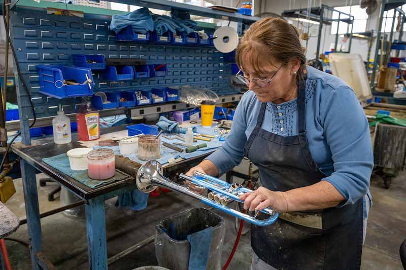 A woman works on a trumpet in on a factory floor. A work bench next to her is full of tools and other supplies.