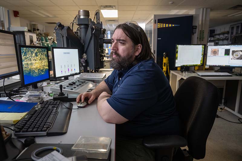 A man with long hair and a beard looks at computer screens in a laboratory.