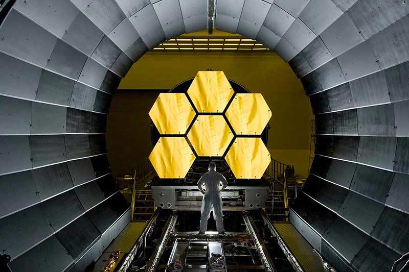 A person in a hazmat suit stands with hands-on-hips in front of gold hexagons part of the Webb telescope.