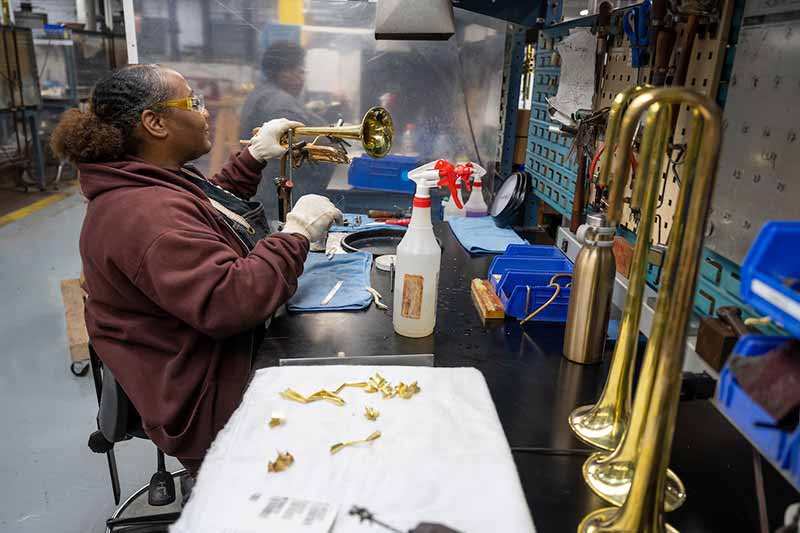 A Black woman wearing eye protection and gloves sits at a table and solders onto a trumpet.