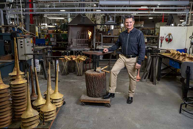 A man stands in a brass instrument factory next to stacks of unfinished bells for trumpets.