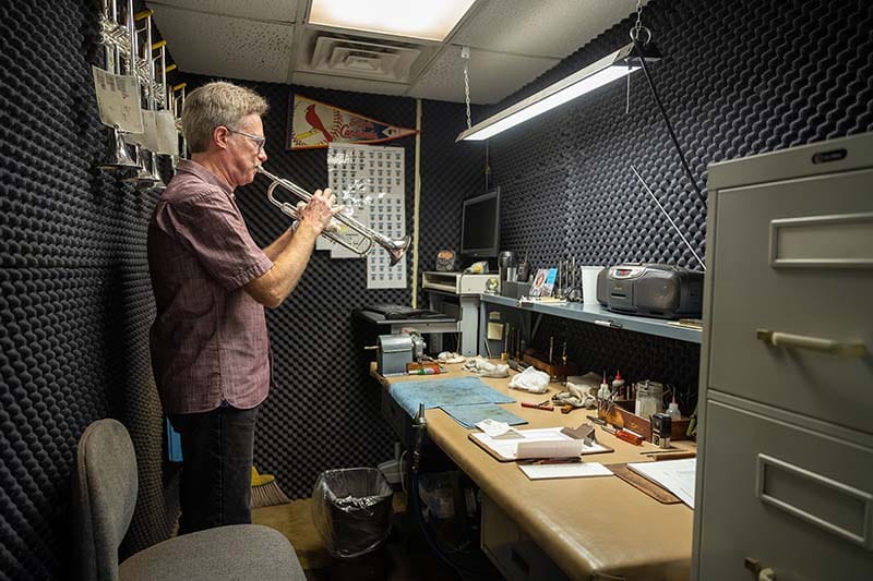 A man plays a trumpet in a soundproofed room with a desk.