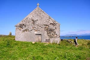 A man walks beside an ancient monastic building. Green grasses, blue skies.