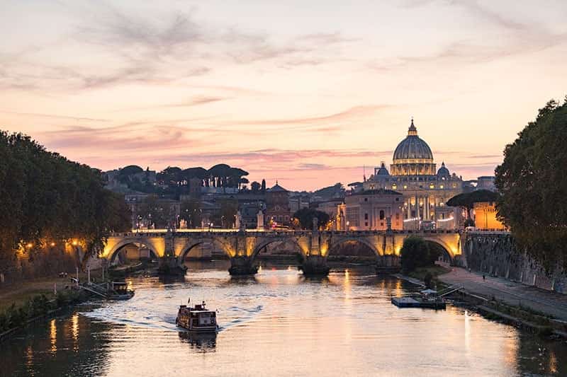 Sun sets over the city of Rome. In the foreground is the Sant'Angelo bridge which spans the Tiber River. In the background to the right is the dome of  Saint Peter's Basilica.