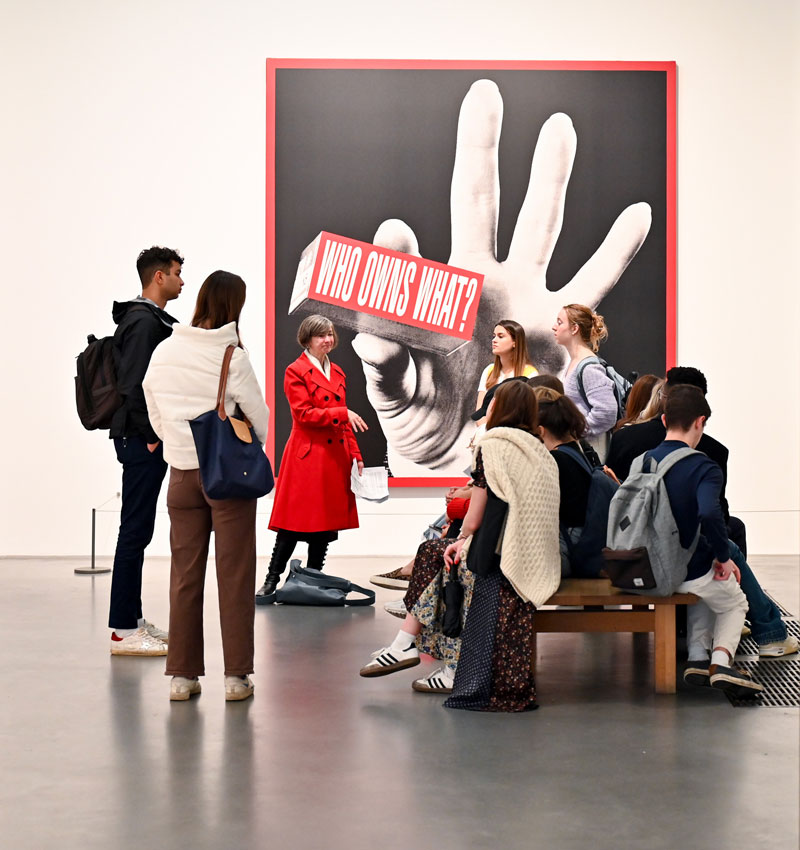 Lois Oliver, in a long red coat, talks about artwork with her students. The art is a photograph of a hand holding a sign that says 'Who Owns What?'
