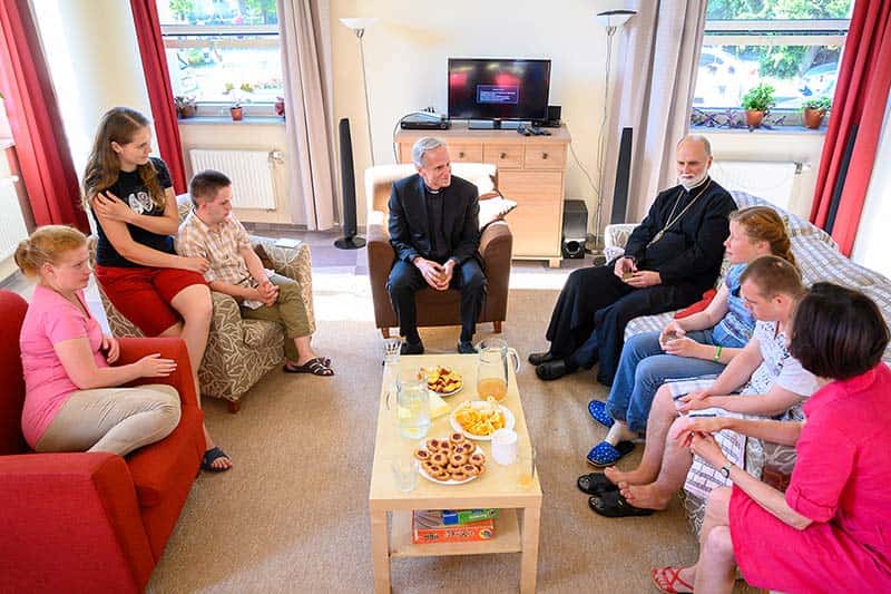 People sit on couches and chairs in a living room, there are pastries, fruit, and juice on a coffee table.