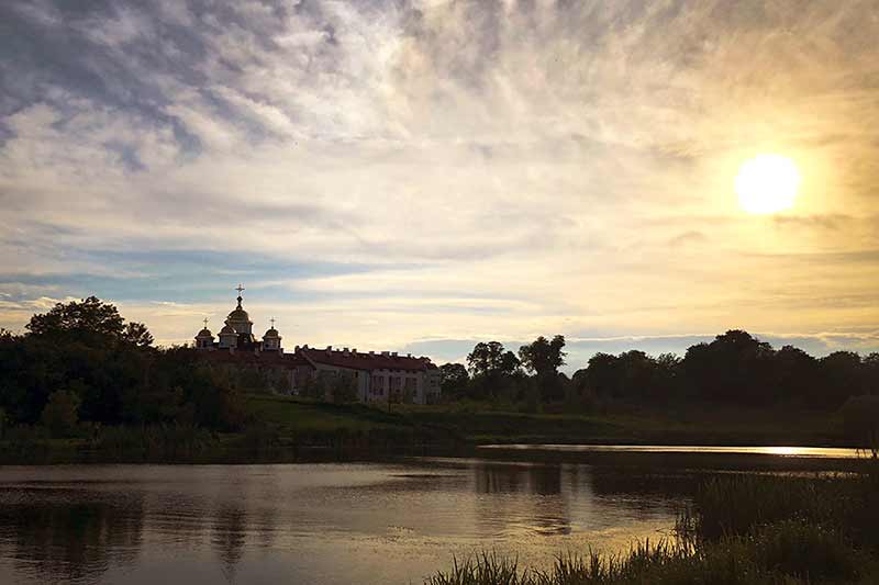 A seminary off in the distance surrounded by several trees and a lake. The sun rises to the east.