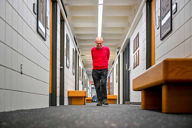 Towards the camera, a man walks down a long, narrow hallway looking down and holding his arms behind his back.