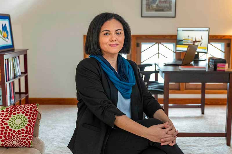 A woman sits on a stool in a home office with her legs crossed and her hands holding her knee.