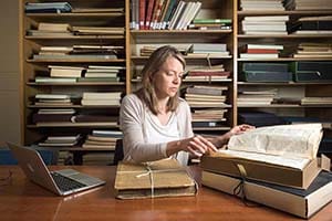 A female scholar sits at a desk with a pile of old books and a laptop.