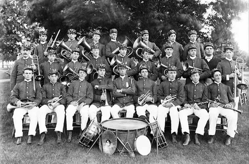 An archival photo of the Notre Dame Marching Band posing for a photo in three rows holding wind instruments. In front of the group lays drums, a tambourine and a cymbal.