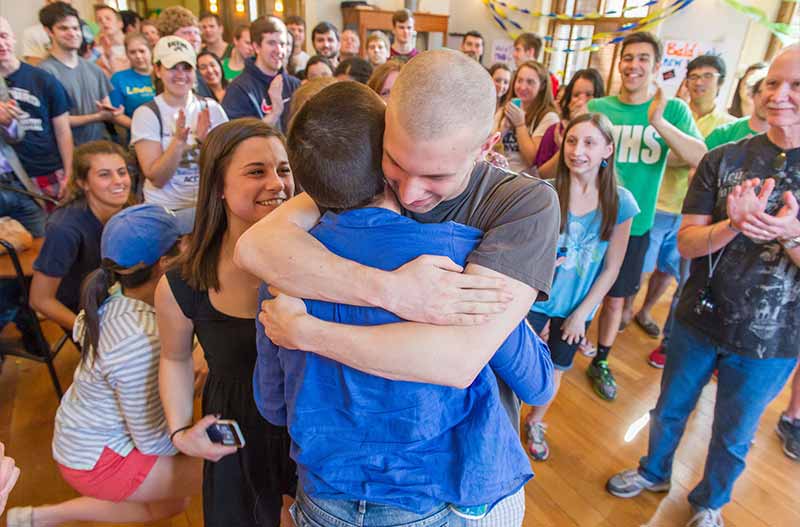 Two people that just shaved their heads hug each other in front of a large group of people.