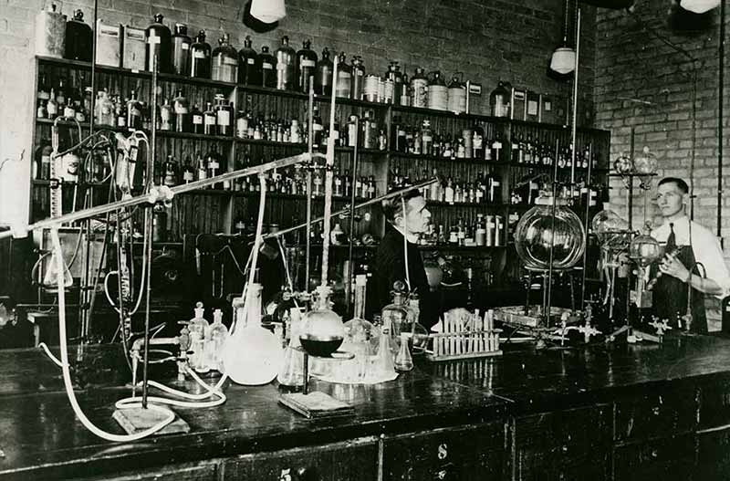 An archival photo of a priest in a chemistry laboratory with a student standing behind a large table with beakers and other lab equipment. There is a shelf that takes up most of the back wall with with different sizes of bottles.