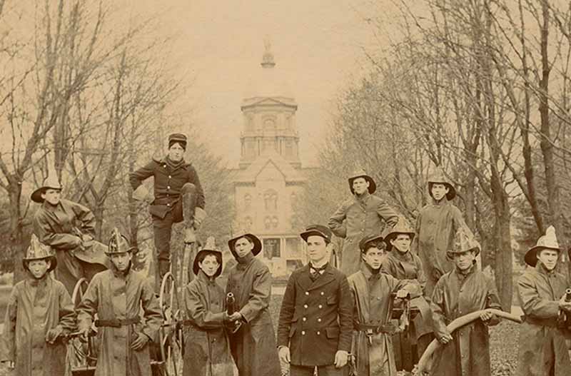 An archival photo of the Notre Dame Fire Department firemen, wearing coats and helmets and some holding onto a hose, posed on Main Quad in front of the Main Building.